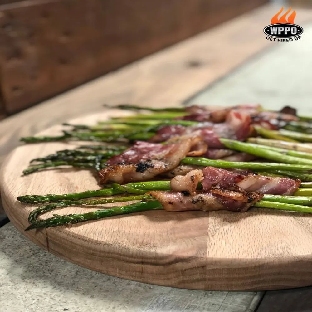 Bacon-wrapped asparagus cooked in WPPO Lil Luigi portable wood-fired oven, served on wooden cutting board.