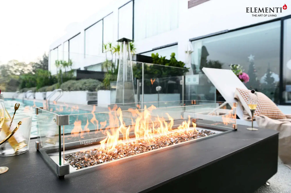 A close-up shot of the fire pit's linear burner, showing the clear glass wind guard surrounding the flames and shimmering bronze glass rocks.