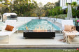 A sleek, black rectangular fire pit table with a marble-textured top centered between two white lounge chairs. The fire is lit with orange flames dancing over glass rocks, situated on a patio overlooking a turquoise swimming pool.