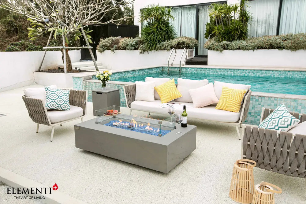High-end backyard patio featuring the Meteora rectangular gas fire pit table as a centerpiece, paired with a designer woven chair and a classic silver trailer.