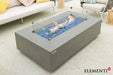 Close-up of the Meteora fire table’s linear burner, highlighting the clear tempered glass wind guard protecting a warm flame over natural lava rocks.