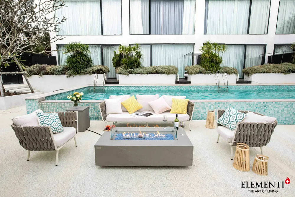 Elementi Plus OFG410SG Meteora 60,000 BTU fire table with a handcrafted slate grey concrete finish, styled on a luxury poolside patio with modern white lounge seating.