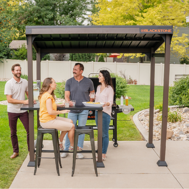 Outdoor gathering around Blackstone 5'x8' Pavilion with grilled food, stools, and built-in counters