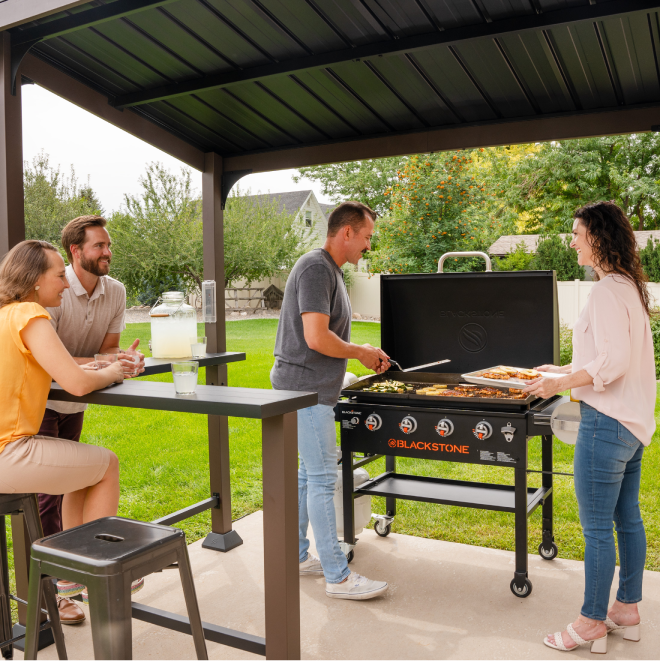 Friends cooking and dining under the Blackstone 5'x8' Outdoor Griddle Pavilion with bartop counters and griddle setup