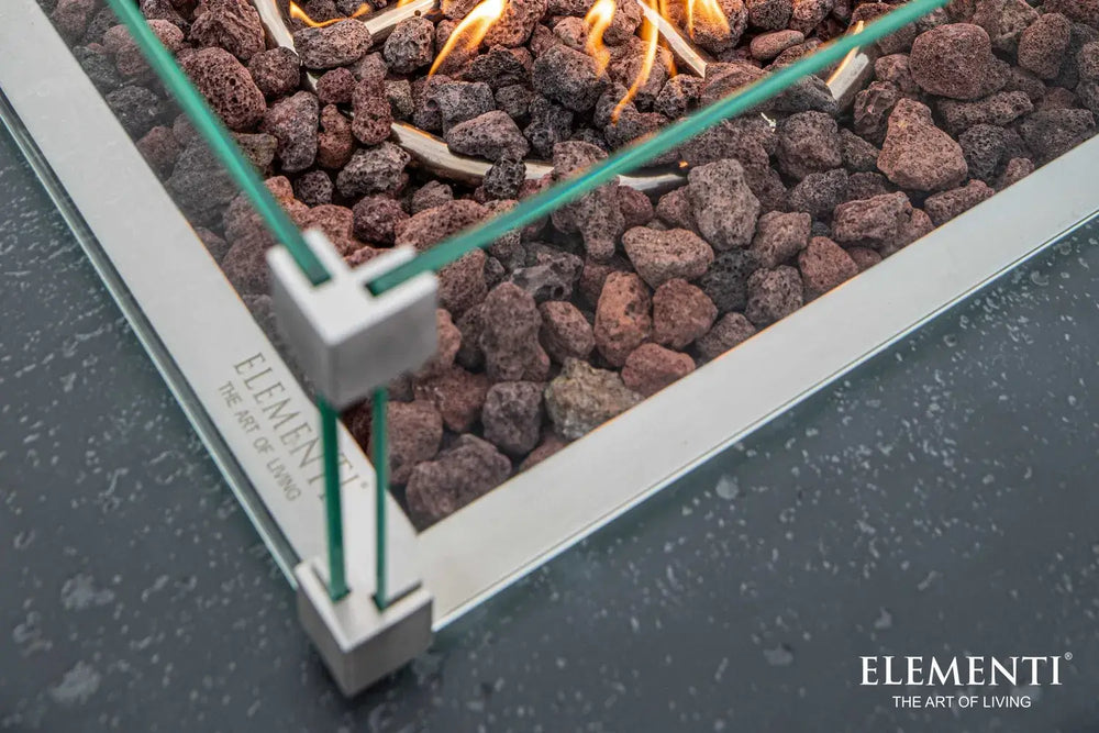 Close-up detail of the Manhattan fire table's stainless steel burner, lava rocks, and glass wind guard corner bracket.