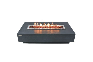 Lit Elementi Hampton OFG139DG Fire Table 56" Dark Gray with glass wind guard and lava rocks on white.