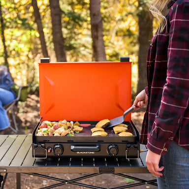 Woman grilling potatoes and veggies on a Blackstone portable camping griddle in a forest setting, showcasing easy-to-use controls and even heat distribution.