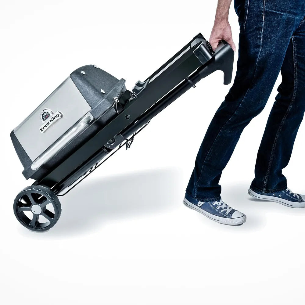 Broil King Porta-Chef AT220 portable gas grill folded into transport mode, being wheeled by a person using the integrated handle and large black wheels.