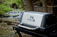 Close-up of the Broil King Porta-Chef AT220 stainless steel lid and thermometer, mounted on the portable black cart in an outdoor wooded setting near a stream.
