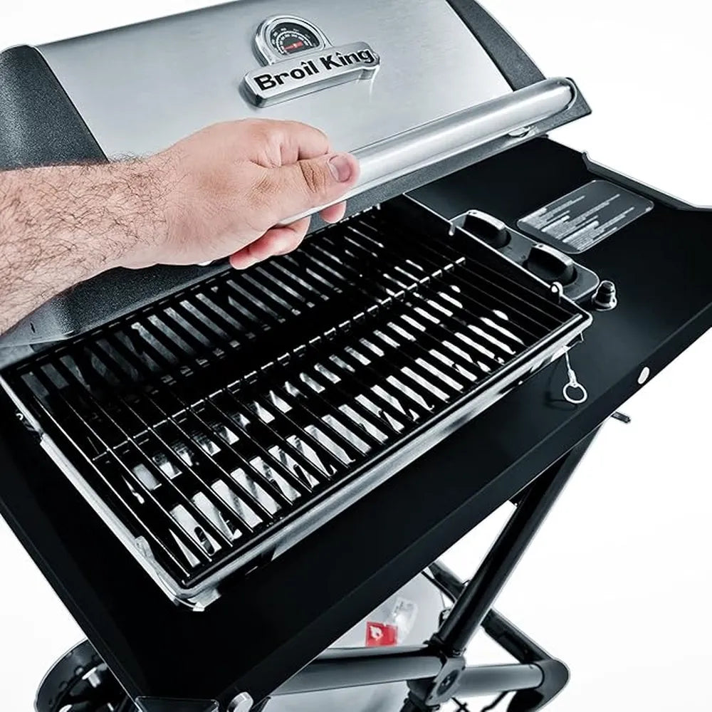 Close-up of a hand lifting the lid on the Broil King Porta-Chef AT220 portable gas grill, showing the stainless steel hood, cooking grates, and side shelf details.