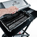 Close-up of a hand lifting the lid on the Broil King Porta-Chef AT220 portable gas grill, showing the stainless steel hood, cooking grates, and side shelf details.