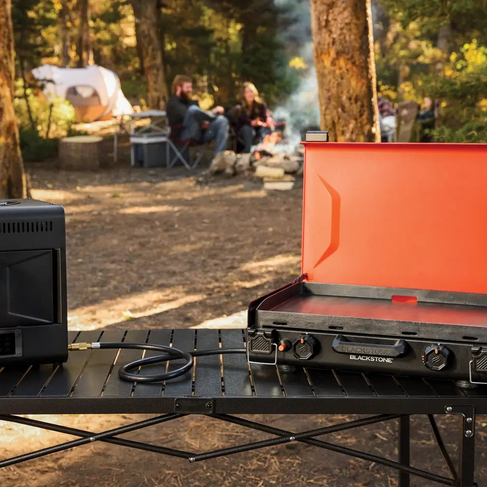 Blackstone 20"x14" griddle connected to a propane tank on a camp table in a forest campsite, ideal for tailgating and camping meals.
