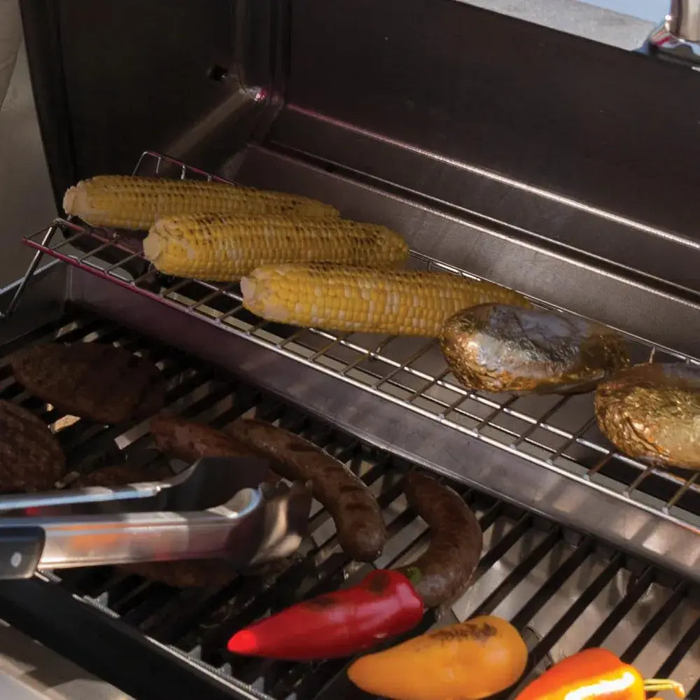 Close-up inside the Broil King Porta-Chef 320 grill showing warming rack with corn and potatoes above the main grate cooking sausages, burgers, and peppers.