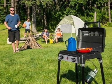 Broil King Porta-Chef Stove 200 portable propane stove in use at a campsite, cooking outdoors near a tent and campfire with people relaxing in the background.