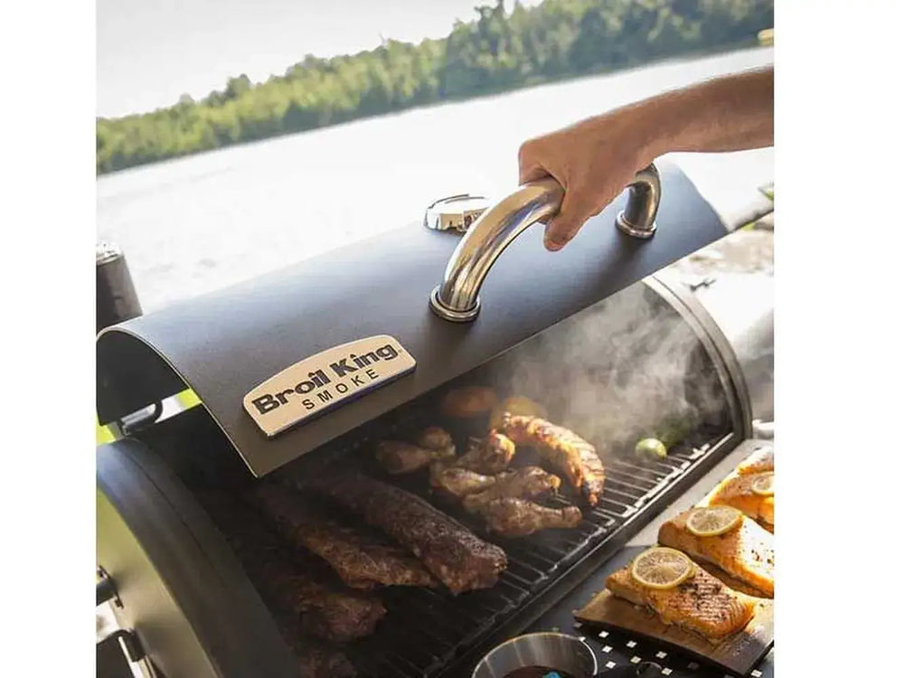 Hand lifting the lid of the Broil King Regal Charcoal Grill 400 as meat smokes on the grill beside cedar plank salmon.