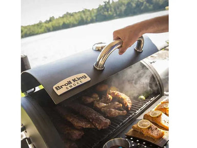 Hand lifting the lid of the Broil King Regal Charcoal Grill 400 as meat smokes on the grill beside cedar plank salmon.
