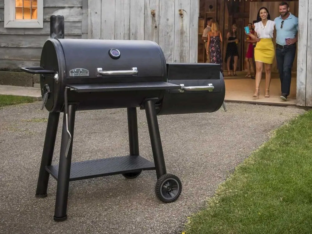 Broil King Regal Charcoal Offset 500 smoker grill displayed outdoors on a patio in front of a rustic building, with guests walking out in the background.