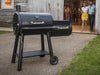 Broil King Regal Charcoal Offset 500 smoker grill displayed outdoors on a patio in front of a rustic building, with guests walking out in the background.