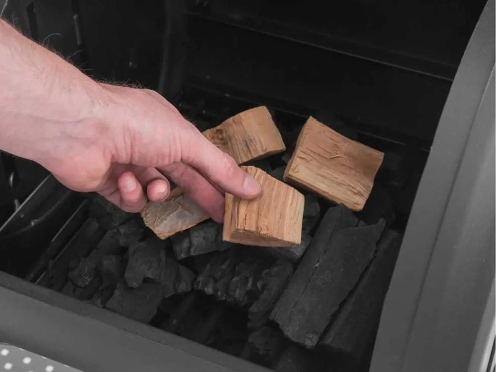 Hand placing wood chunks over charcoal inside the Broil King Regal Charcoal Offset 500 side firebox for smoking flavor.