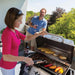 Woman grilling on a Broil King Porta-Chef 320 portable gas grill on a balcony, cooking corn, potatoes, sausages, burgers, and peppers while another person stands nearby with a glass of wine.