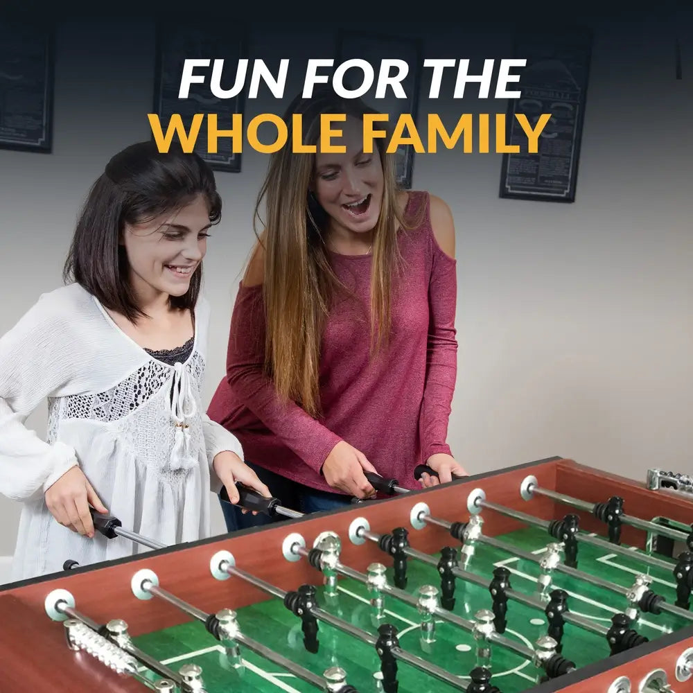 An image titled "Fun For The Whole Family" showing two young women smiling and playing with the Hathaway Metropolitan 54-inch Foosball Table in a game room setting.