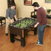 Two men playing foosball on the Hathaway Primo 56-inch table in a cozy game room with wooden flooring and framed wall art.