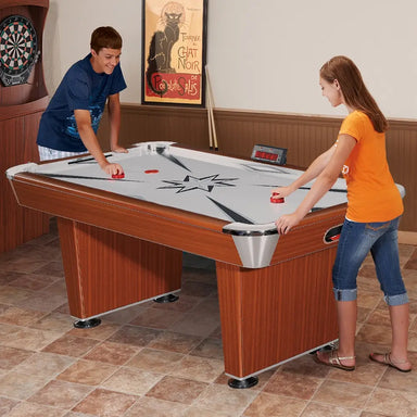 Two teenagers playing on a Hathaway air hockey table in a game room setting. The table features a glossy white playfield, black and gray star graphics, and a digital scoreboard. Dartboard and framed artwork in the background.