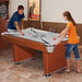 Two teenagers playing on a Hathaway air hockey table in a game room setting. The table features a glossy white playfield, black and gray star graphics, and a digital scoreboard. Dartboard and framed artwork in the background.