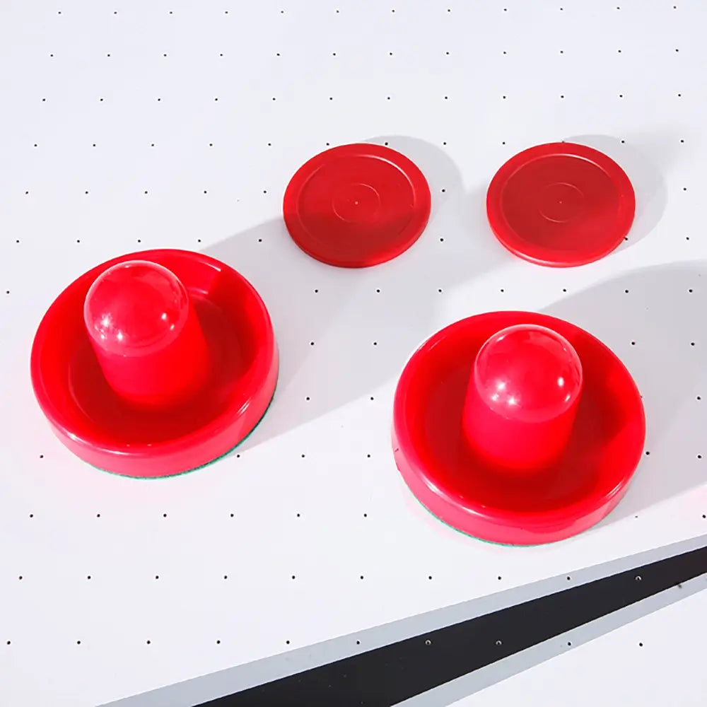 Close-up of two red air hockey pushers and two red pucks placed on the white playfield with micro-perforations for air flow. Essential accessories for playing air hockey.
