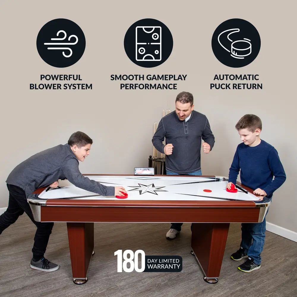 Family playing on a Hathaway air hockey table, with overlay icons indicating key features: powerful blower system, smooth gameplay performance, and automatic puck return. Image promotes 180-day limited warranty. Table shown with cherry wood cabinet, white playfield, and digital scoreboard.