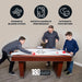 Family playing on a Hathaway air hockey table, with overlay icons indicating key features: powerful blower system, smooth gameplay performance, and automatic puck return. Image promotes 180-day limited warranty. Table shown with cherry wood cabinet, white playfield, and digital scoreboard.