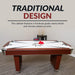 Hathaway air hockey table shown in a modern game room, emphasizing its traditional design with furniture-grade cherry finish and chrome-plated trim. Red pushers and pucks rest on a white playfield with black star graphic. Cue stand and accessories visible in background.