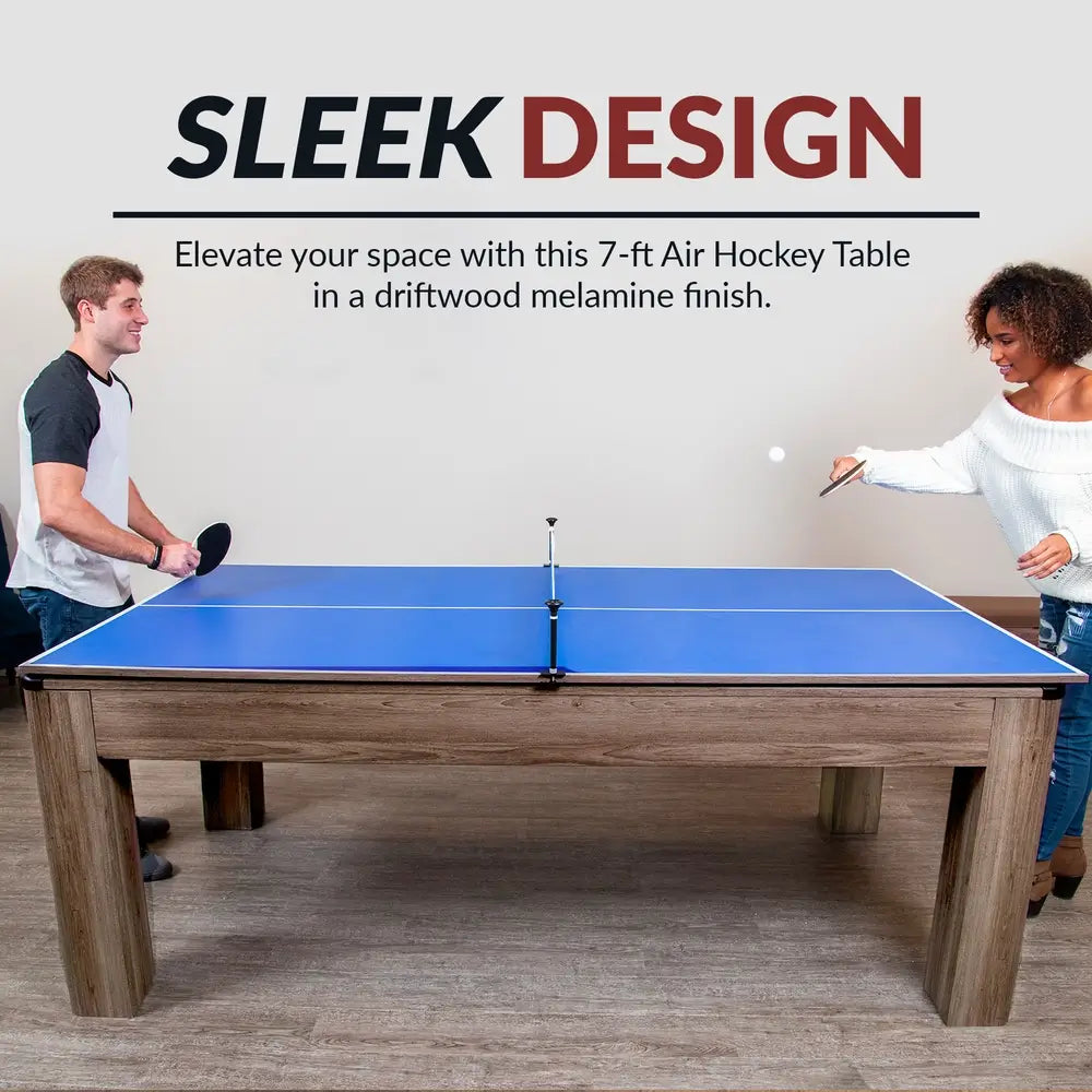 Modern 7-foot air hockey and ping pong table with driftwood melamine finish, shown in table tennis mode with two players enjoying a game.
