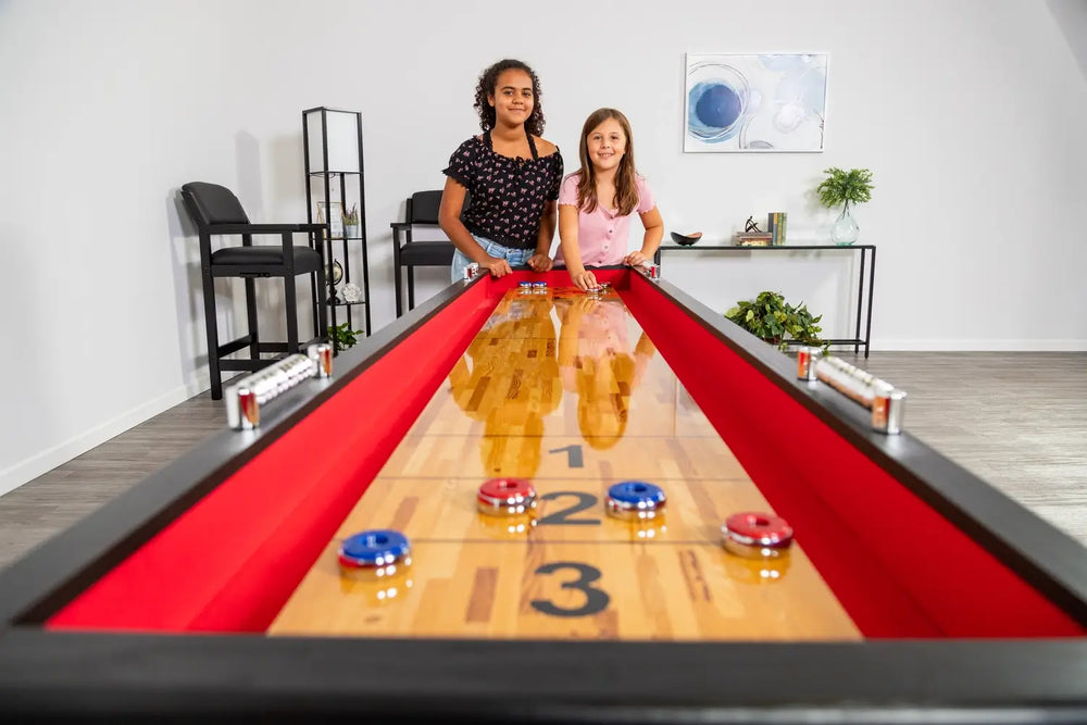 Two kids playing shuffleboard on a Hathaway table with red padded gutters and chrome scoring units in a modern game room.