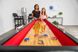 Two kids playing shuffleboard on a Hathaway table with red padded gutters and chrome scoring units in a modern game room.
