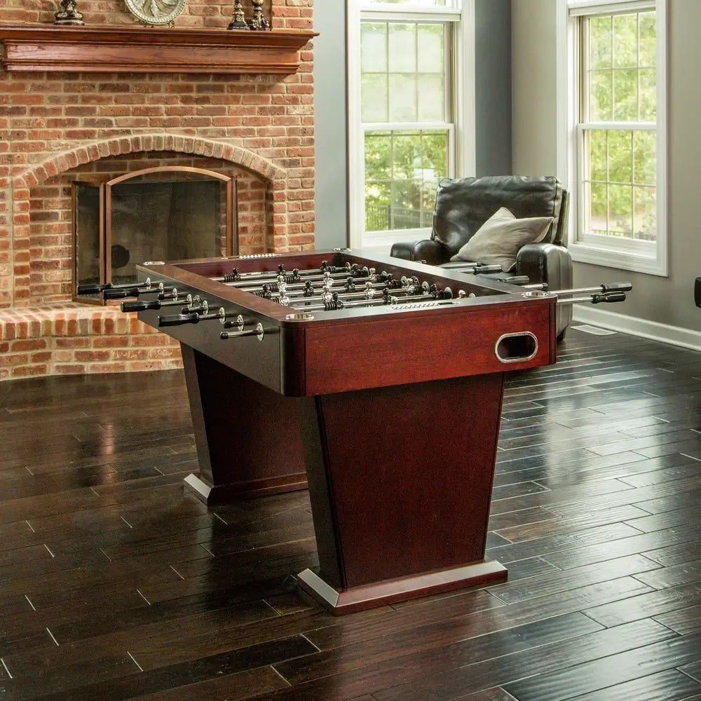 Espresso-finish foosball table with chrome accents and modern pedestal legs, staged in an elegant game room with brick fireplace and leather chair.