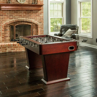 Espresso-finish foosball table with chrome accents and modern pedestal legs, staged in an elegant game room with brick fireplace and leather chair.