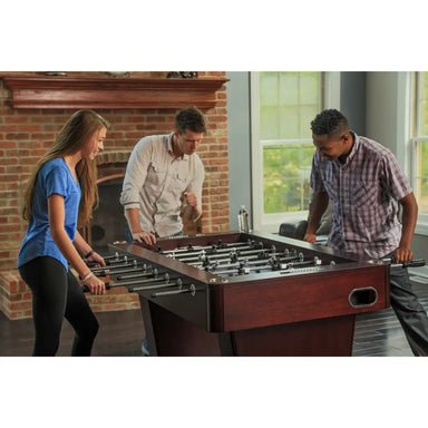 Three people playing an intense foosball match on a modern espresso-finish table in a stylish living space with natural light and brick accents.