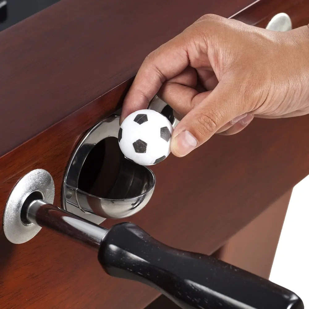 Hand retrieving foosball from chrome-finished ball return slot on side of espresso-wood foosball table, highlighting accessible gameplay design.