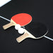 Red and black table tennis paddles with two white ping pong balls placed on matte black table tennis surface with center line.