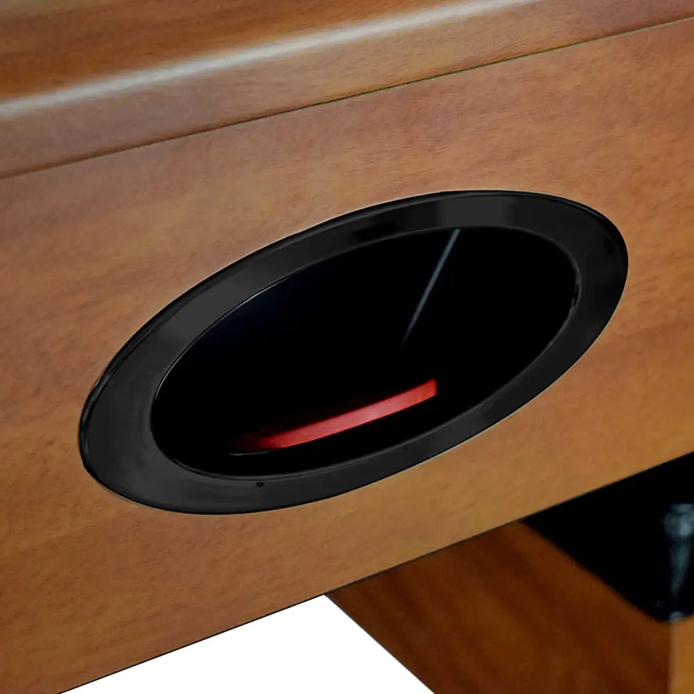 Detail view of puck retrieval goal slot with black trim on wooden air hockey table, showing red puck inside goal compartment.