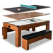 Exploded view of multi-functional game table showing removable tops for table tennis, dining, and air hockey; features cherry wood frame, white playfield, and black cushioned bench seating.