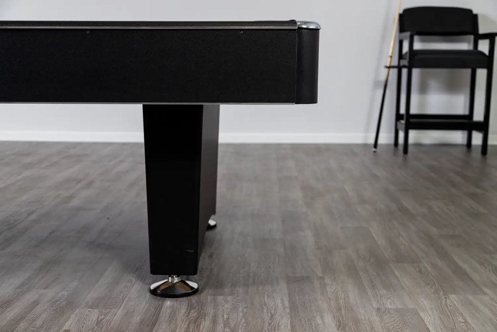 Side profile of the Hathaway Hustler pool table showing black pedestal-style leg, textured black cabinet finish, chrome leg levelers, and gray wood-look flooring with pool cues and chairs in the background.