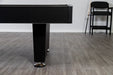 Side profile of the Hathaway Hustler pool table showing black pedestal-style leg, textured black cabinet finish, chrome leg levelers, and gray wood-look flooring with pool cues and chairs in the background.