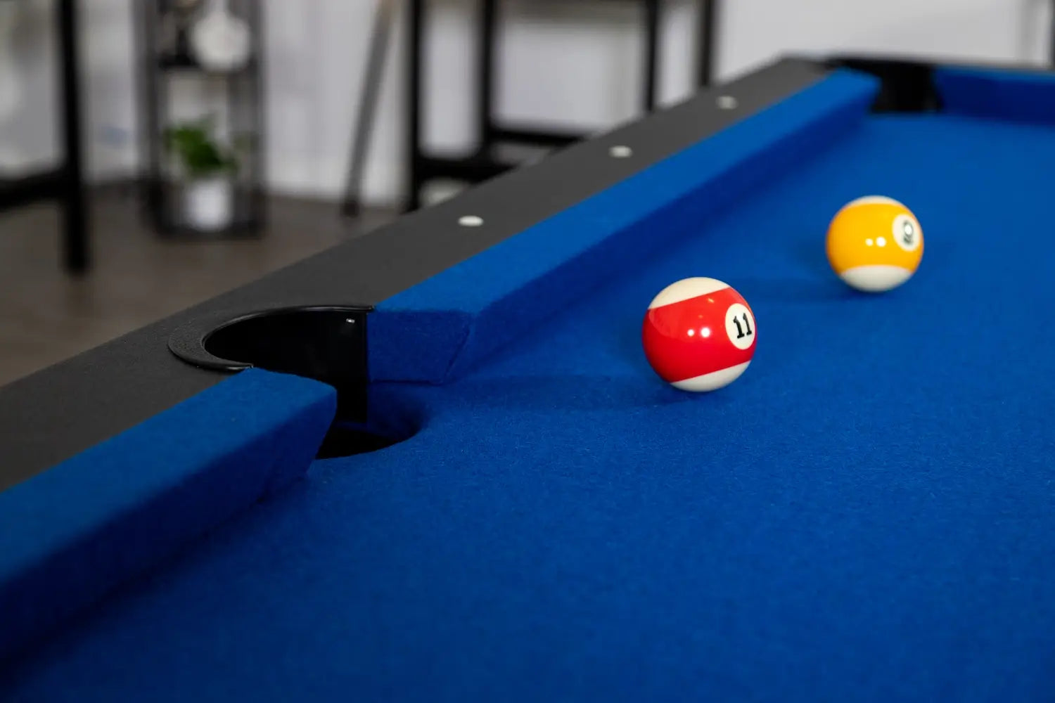 Close-up of two billiard balls (#11 red stripe and #9 yellow stripe) on blue wool-blend felt near a corner pocket with black plastic liner and black top rail on the Hathaway Hustler pool table.