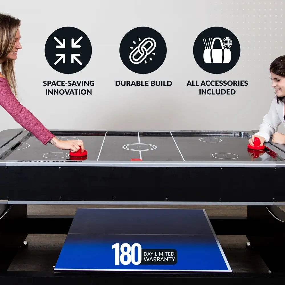 Two people playing air hockey on a black multi-game table with space-saving innovation, durable build, and all accessories included; includes 180-day limited warranty.