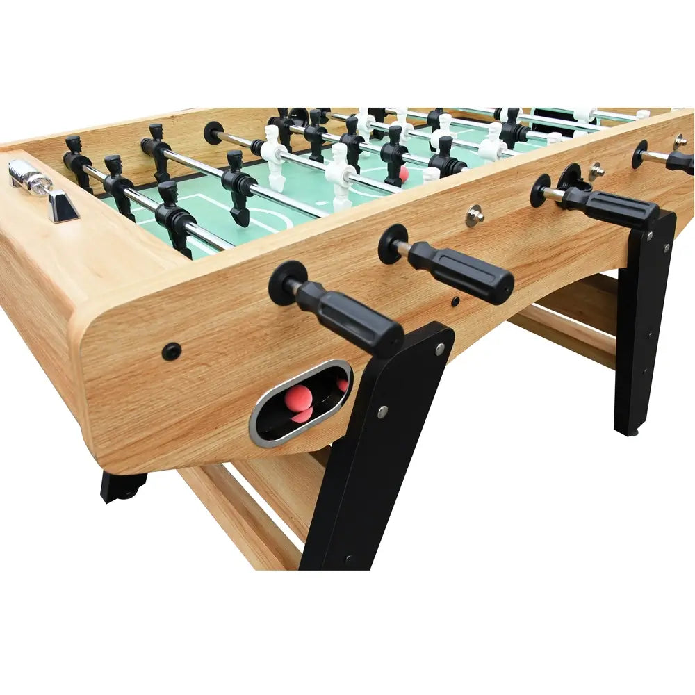 Close-up of foosball table corner showing black ball return slot with red balls, ergonomic black handles, and light wood cabinet finish.