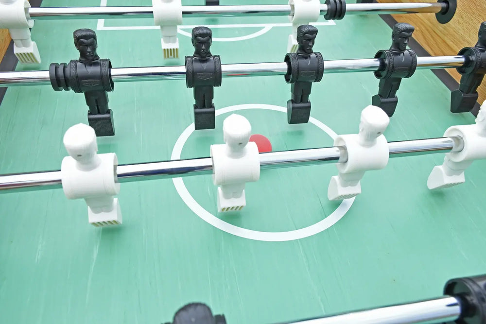 Close-up of foosball players in black and white on chrome rods, with red ball positioned in center circle of a green playfield.