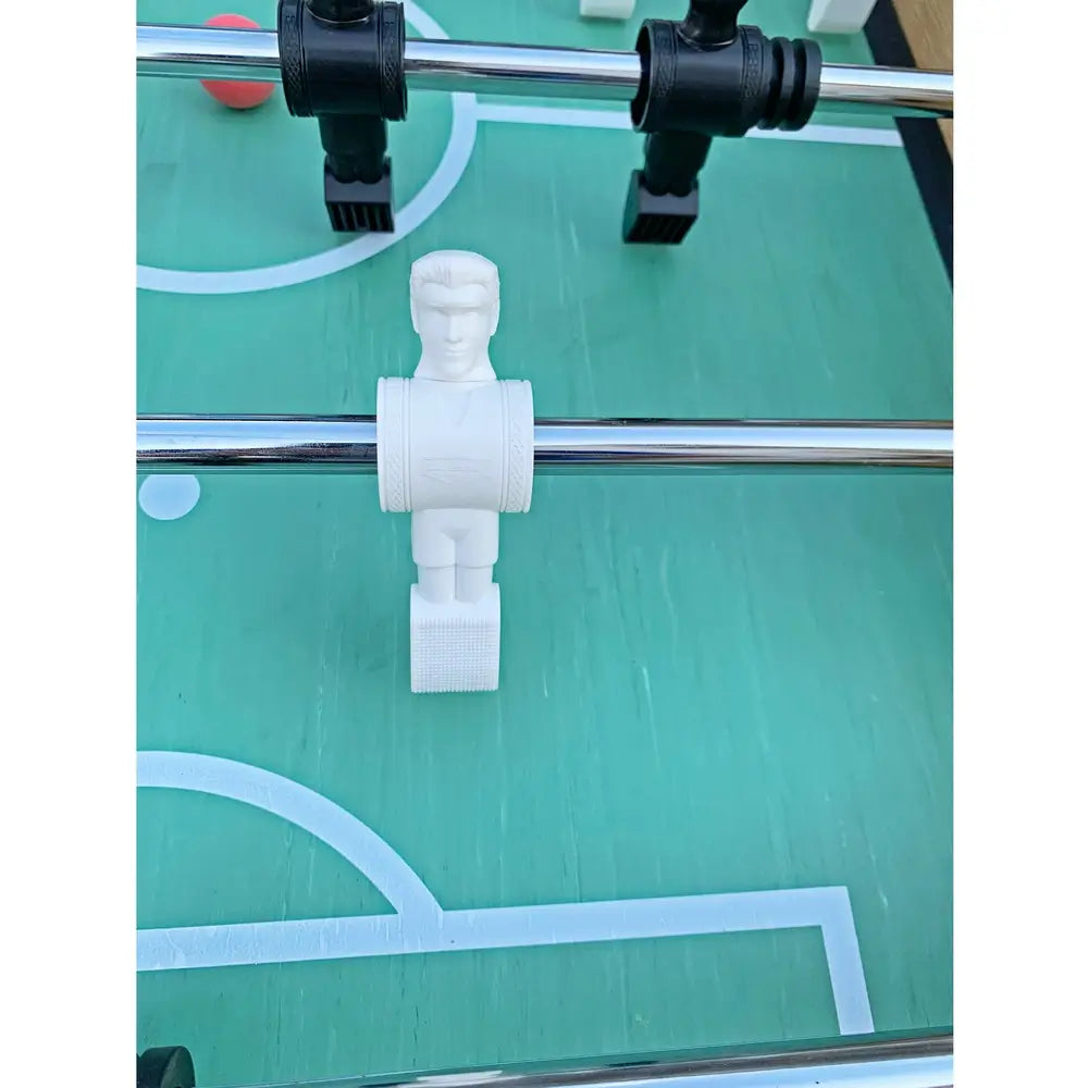 Zoom-in of white foosball player figure with facial features and textured feet, mounted on chrome rod above green field with goal area lines.