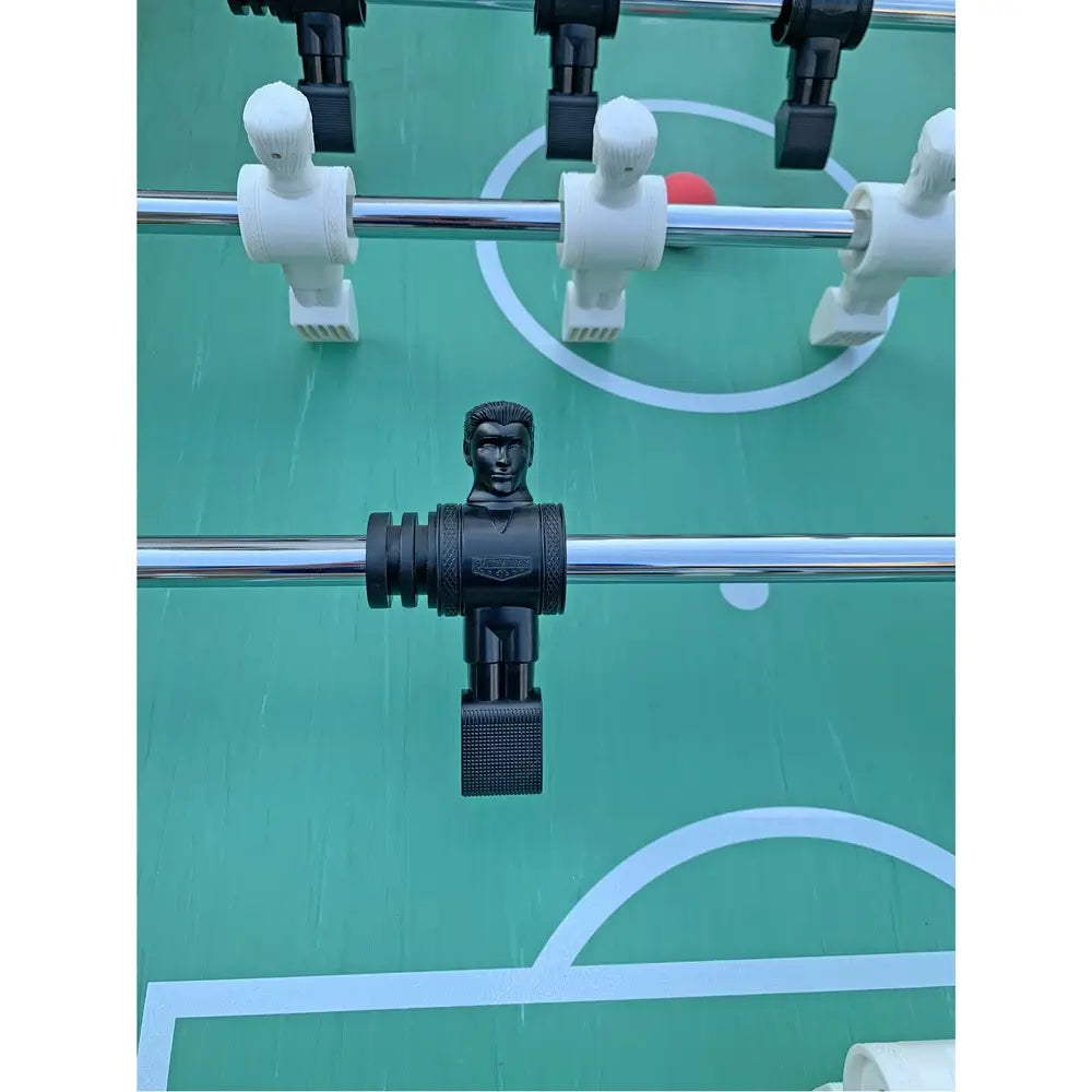 Close-up of black foosball player with defined facial features and textured foot, mounted on chrome rod above soccer field-style surface.